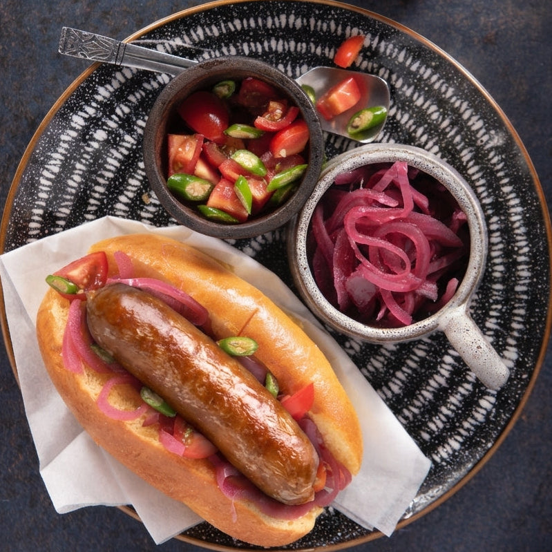 Hot dog with beef sausage on a plate with side dishes of tomatoes and red onions.